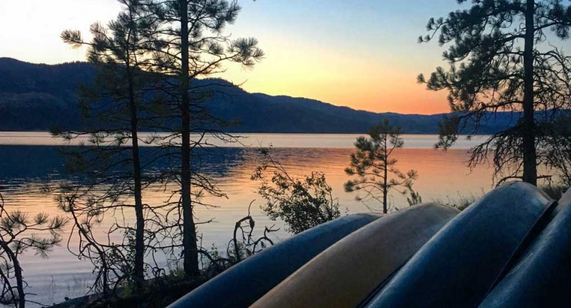 a group of canoes rest on the shore beside a calm lake while the sun is setting